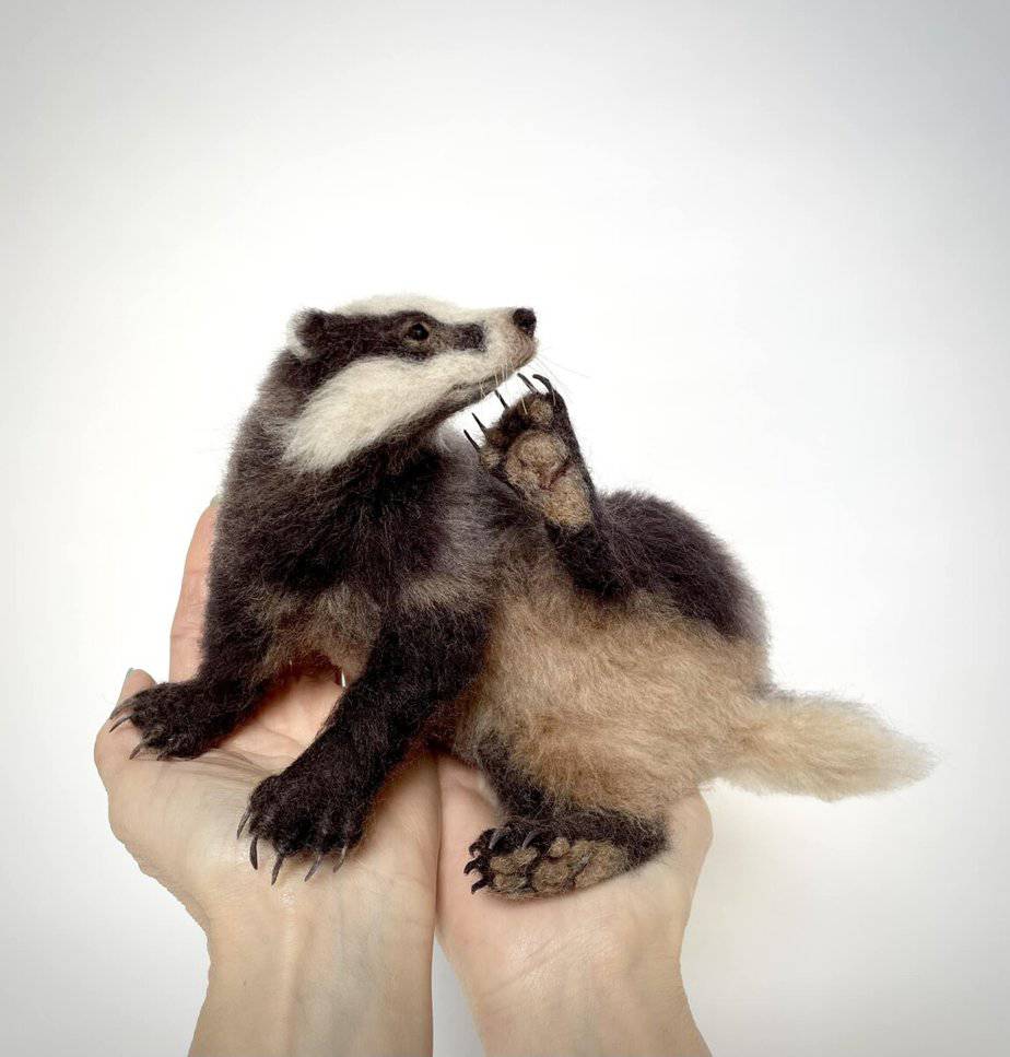 Two hands holding a small lifelike needle-felted badger sculpture against a plain white background.