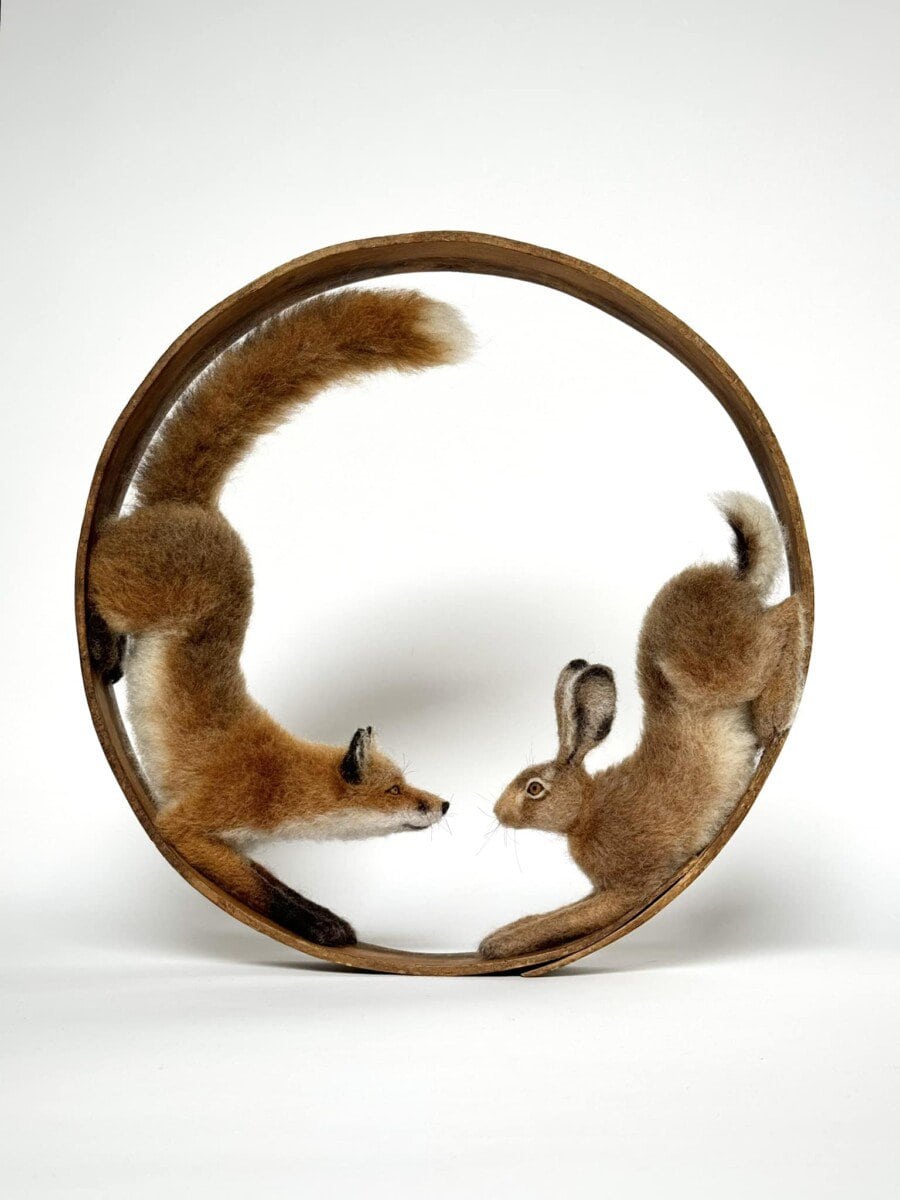 Wool sculptures of a fox and a rabbit facing each other inside a circular wooden frame on a white background.