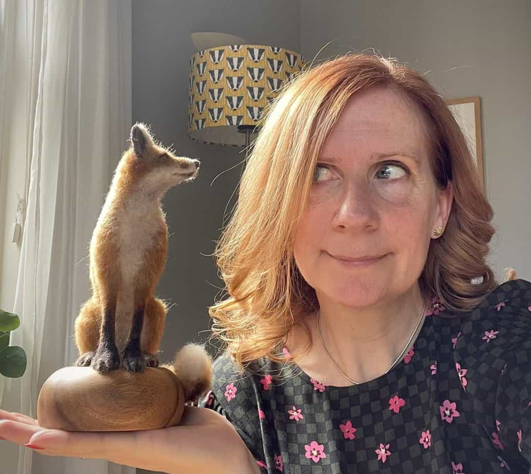 Woman with red hair, holding a small faux fox sculpture in her hand, looking up at it with a playful expression.