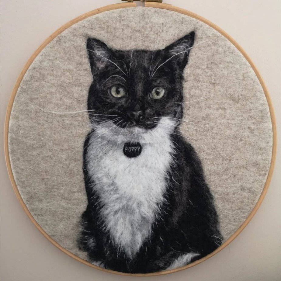 Embroidery hoop art depicting a black and white cat with a tag labeled "Poppy" on a beige background.