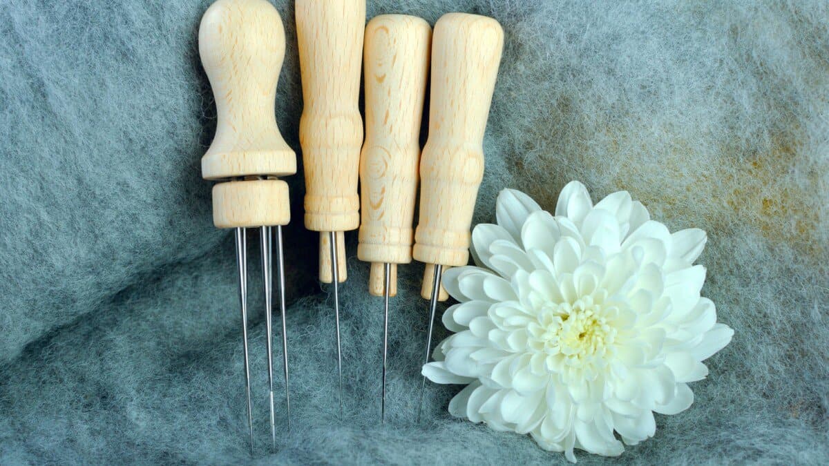 Wood-handled wool felting needles of various sizes arranged on grey felt, alongside a white chrysanthemum flower.