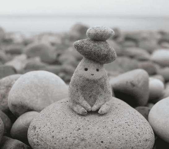 Small rock sculpture shaped like a creature with a pebble hat, sitting on a larger rock among many rounded stones on a beach.