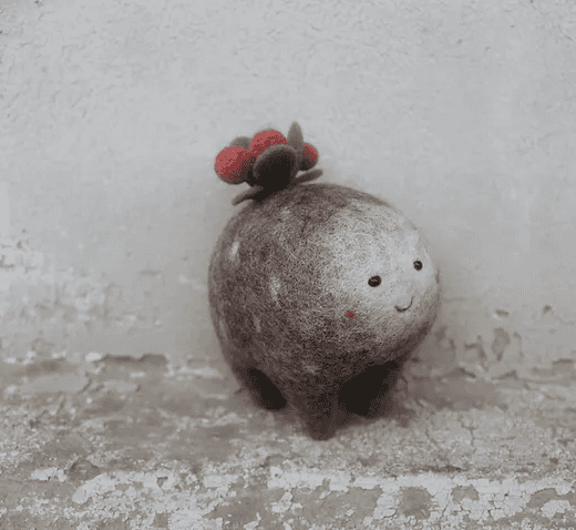 A small, round felt creature with a smiling face and red berries on its head, sitting on a textured, weathered surface.