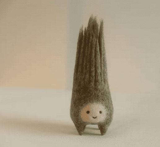 A small, smiling character with a round face and tall, textured, cone-shaped hair standing on a light surface.