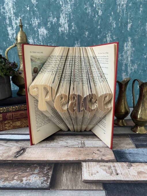 Open book with folded pages spelling "Peace" on a wooden surface, surrounded by antique brass vases and a vintage background.