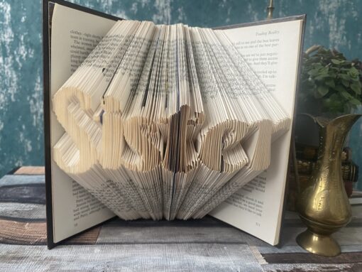 An open book with pages folded to spell "Sister," placed on a wooden table next to a brass vase and a small potted plant.