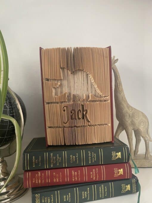 A folded book sculpture depicting a dinosaur and the name "Jack" sits on a stack of hardcover books next to a globe and giraffe statue.