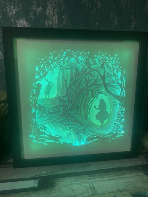 Shadowbox art featuring a green illuminated forest scene with two silhouetted figures, a girl in a dress and a man holding a staff.