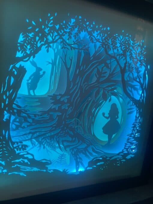 Silhouette of a girl and a rabbit in a forest made from layered paper art, illuminated with blue backlighting.