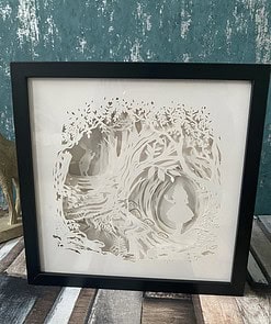 Framed papercut art depicting a girl in a forest surrounded by intricate tree and foliage designs. Plaque is placed on wooden surface.