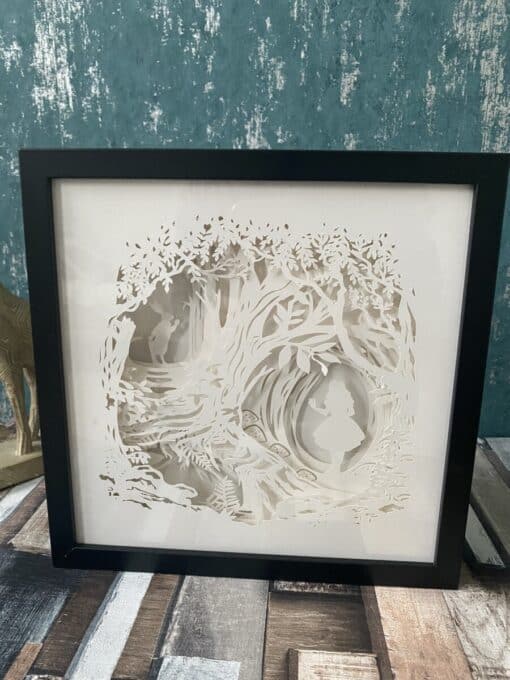 Framed papercut art depicting a girl in a forest surrounded by intricate tree and foliage designs. Plaque is placed on wooden surface.