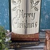 An open book with pages intricately folded to spell "Merry Christmas" and holly designs, placed on top of three closed books.