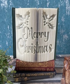 An open book with pages intricately folded to spell "Merry Christmas" and holly designs, placed on top of three closed books.