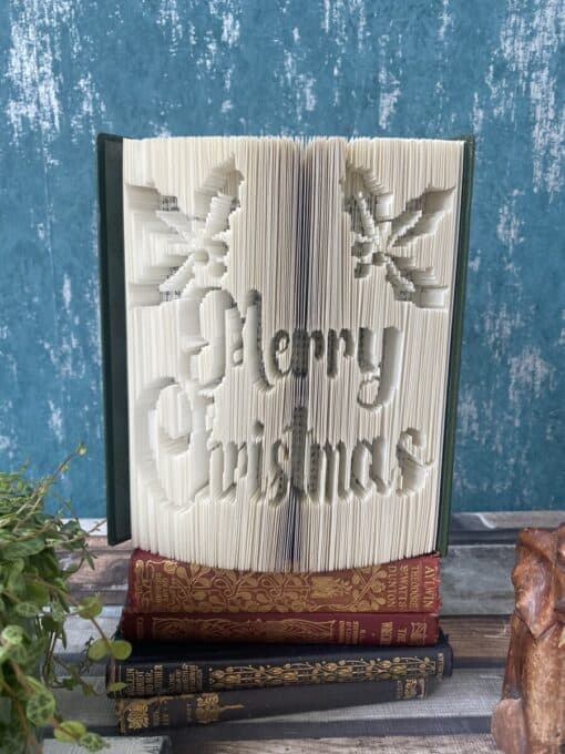 An open book with pages intricately folded to spell "Merry Christmas" and holly designs, placed on top of three closed books.