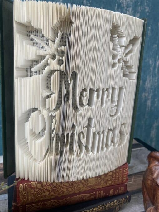 An open book with pages folded to spell "Merry Christmas" and depict two snowflakes, standing on two other books.