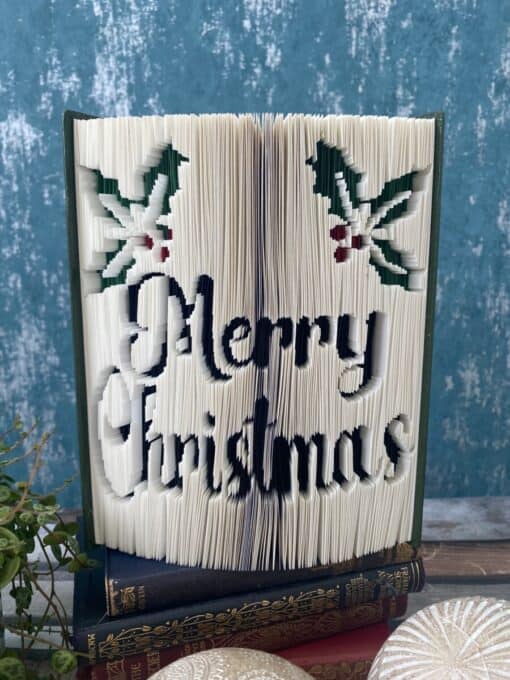 A book with folded pages creating a "Merry Christmas" design, decorated with holly and berries, rests on a table.