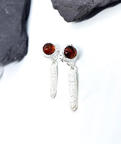 Silver drop earrings with textured long dangles and garnet-coloured stone studs, placed on a white surface with dark stones.