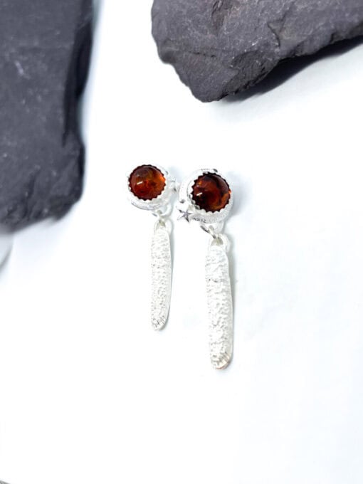 Silver drop earrings with textured long dangles and garnet-coloured stone studs, placed on a white surface with dark stones.