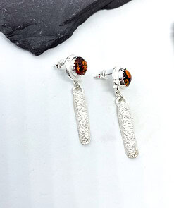 Silver dangle earrings with textured elongated drops and amber stone studs, displayed on a white surface with a slate backdrop.