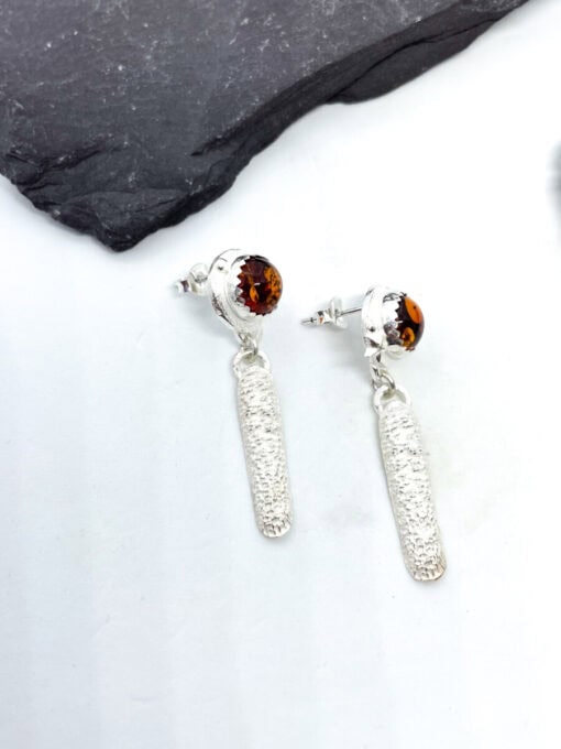 Silver dangle earrings with textured elongated drops and amber stone studs, displayed on a white surface with a slate backdrop.