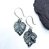 Silver leaf-shaped earrings featuring intricate veins and small crescent moon accents, displayed against a white background with a black stone.