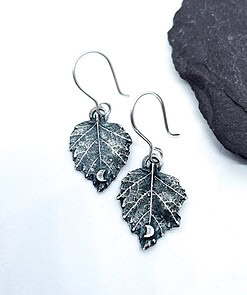 Silver leaf-shaped earrings featuring intricate veins and small crescent moon accents, displayed against a white background with a black stone.