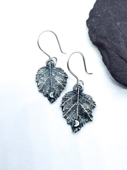 Silver leaf-shaped earrings featuring intricate veins and small crescent moon accents, displayed against a white background with a black stone.