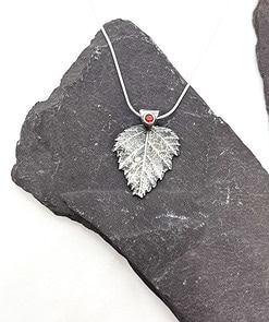 Silver leaf pendant with a red gemstone on a silver chain, displayed on a dark slate background.