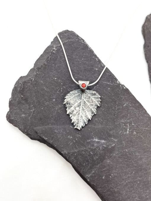 Silver leaf pendant with a red gemstone on a silver chain, displayed on a dark slate background.