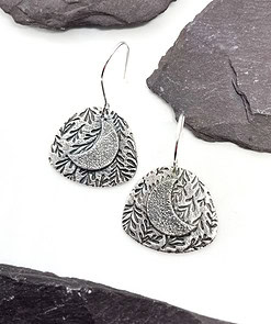 Silver earrings featuring a textured crescent moon and foliage design, placed on a white surface with gray slate accents.