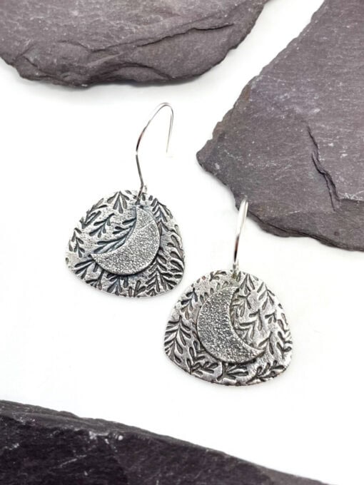 Silver earrings featuring a textured crescent moon and foliage design, placed on a white surface with gray slate accents.