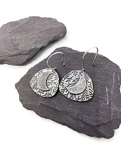 Silver crescent moon earrings with leaf patterns on dark slate stones, showcasing intricate carved details.