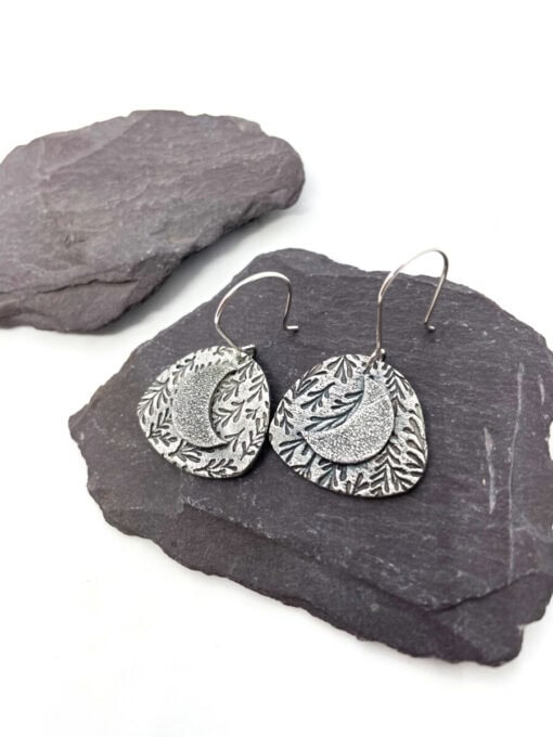 Silver crescent moon earrings with leaf patterns on dark slate stones, showcasing intricate carved details.