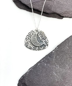 Silver necklace with an engraved pendant featuring a crescent moon and leaf pattern, positioned on a dark slate surface.