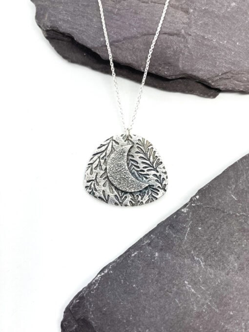 Silver necklace with an engraved pendant featuring a crescent moon and leaf pattern, positioned on a dark slate surface.