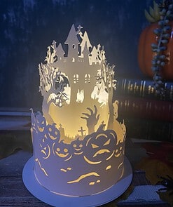 A paper lantern with Halloween-themed cutouts of a haunted house, jack-o'-lanterns, and spooky trees glows in dim light.