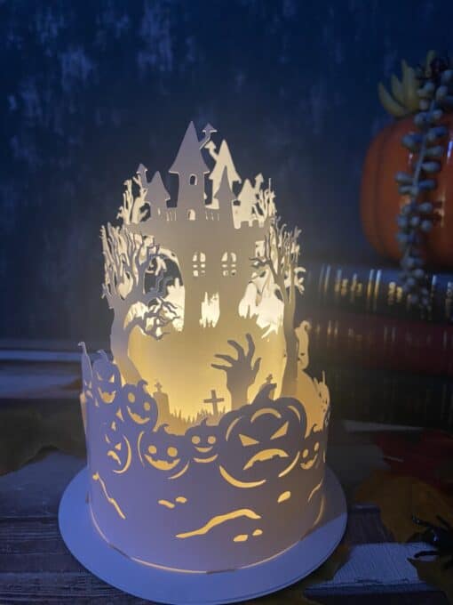 A paper lantern with Halloween-themed cutouts of a haunted house, jack-o'-lanterns, and spooky trees glows in dim light.