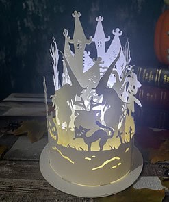 A Halloween-themed paper lantern with witch silhouettes, a black cat, ghostly figures, and a pumpkin, illuminated in the dark.