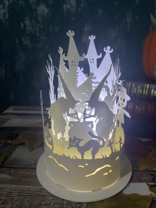 A Halloween-themed paper lantern with witch silhouettes, a black cat, ghostly figures, and a pumpkin, illuminated in the dark.