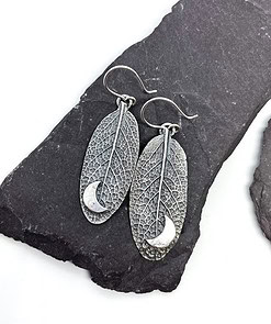 Silver leaf-shaped earrings with intricate vein patterns and crescent moon details, displayed on dark stone slabs.