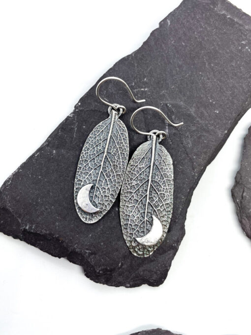 Silver leaf-shaped earrings with intricate vein patterns and crescent moon details, displayed on dark stone slabs.