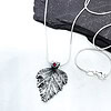 Silver leaf-shaped pendant necklace with a red gemstone on a white background, resting on a dark slate stone.