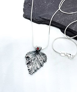 Silver leaf-shaped pendant necklace with a red gemstone on a white background, resting on a dark slate stone.