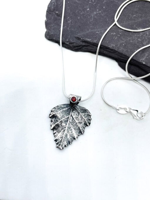 Silver leaf-shaped pendant necklace with a red gemstone on a white background, resting on a dark slate stone.