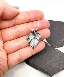 Hand holding a silver leaf-shaped pendant with a red gemstone, displayed on a textured dark slate background.