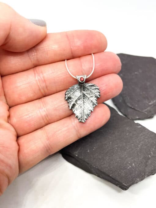 Hand holding a silver leaf-shaped pendant with a red gemstone, displayed on a textured dark slate background.