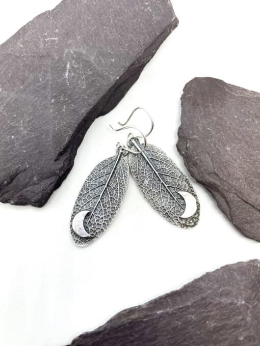 Silver leaf-shaped earrings with intricate vein details and a crescent moon pendant, displayed on dark gray stones.