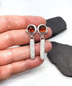 Hand holding silver earrings with round amber stones and star accents on a white background with dark stones.