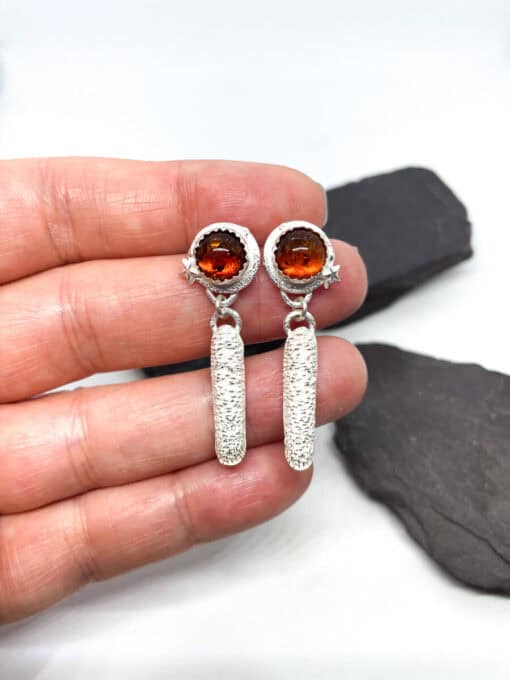 Hand holding silver earrings with round amber stones and star accents on a white background with dark stones.