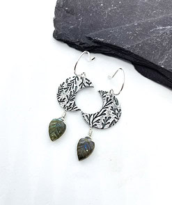 Silver earrings with circular textured design and leaf-shaped green stone pendants, placed on a gray slate slab.